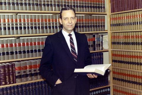 Frank Denholm standing in his law office holding a book. There are shelves of law books behind him.