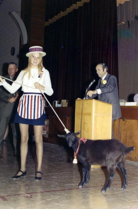 Frank Denholm at a podium speaking to an audience in an auditorium. A woman with a goat on a leash is standing in front of him.