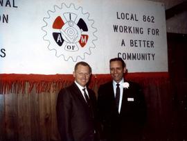Frank Denholm at an International Association of Machinists event. He is standing next to a man in front of a Local 862 sign.
