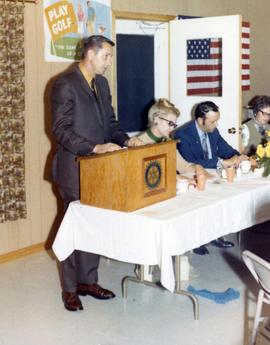 Richard Kneip is speaking to a Rotary Interational gathering. Frank Denholm is seated at the table to the left of Kneip.