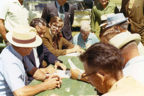 Frank Denholm seated at a picnic table talking with constituents. He is looking at a sheet of paper.