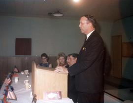 Frank Denholm at podiuim giving a speech at an event. There are people seated to his right.