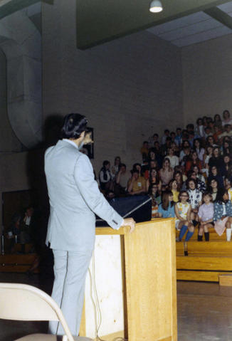 Frank Denholm standing at a podium in a gymnasium giving a talk to an assembly of students in Yankton, South Dakota.