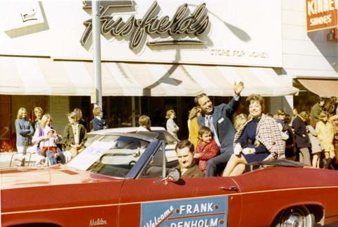 Frank and Millie Denholm riding in a red convertible in the Augustana College Viking Days parade in Sioux Falls, South Dakota. Frank is waving to parade spectators. There is a young girl standing in the back seat next to Frank.