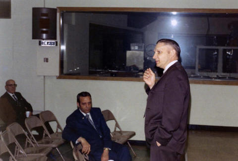 Frank Denholm (seated) at a meeting in a room with a large window. He is listening to a man speak at the front of the room.