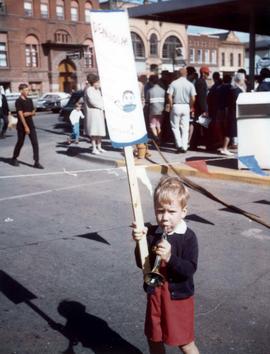 Little boy standing on a street with a Denholm sign in one hand and a toy trumpet in the other hand.