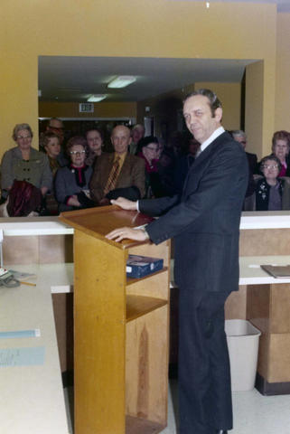 Frank Denholm is standing at a podium behind a desk. A group of people is standing in the hallway.