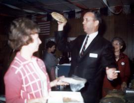 Frank Denholm at a campaign event in Aberdeen, South Dakota. He is holding a pie in his right hand that he is auctioning to the highest bidder. A woman is in the foreground.