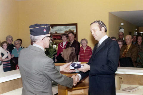 Frank Denholm is at a podium behind a desk. He is shaking the hands with a VFW member and handing him a folded American flag. A group of people is standing in the hallway.